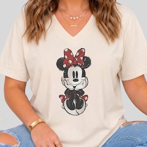 May include: A cream-colored V-neck t-shirt featuring a graphic print of Minnie Mouse. The character is depicted with a red and white polka-dot bow, black ears, and a smiling expression, with her hands resting under her chin in a seated pose.