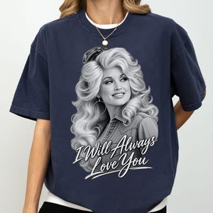 May include: A navy blue short-sleeve t-shirt featuring a black and white graphic print of a woman with voluminous hair wearing a denim jacket. Below the portrait, the text "I Will Always Love You" is printed in a white, stylized script font.