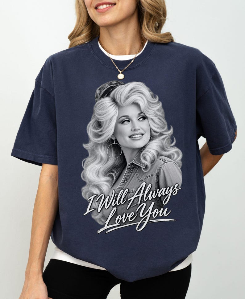 May include: A navy blue short-sleeve t-shirt featuring a black and white graphic print of a woman with voluminous hair wearing a denim jacket. Below the portrait, the text "I Will Always Love You" is printed in a white, stylized script font.