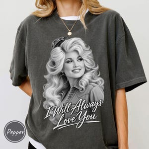 May include: A charcoal gray short-sleeve t-shirt featuring a black and white graphic of a woman with voluminous blonde hair wearing a denim shirt. The text "I Will Always Love You" is printed in white script below the portrait.