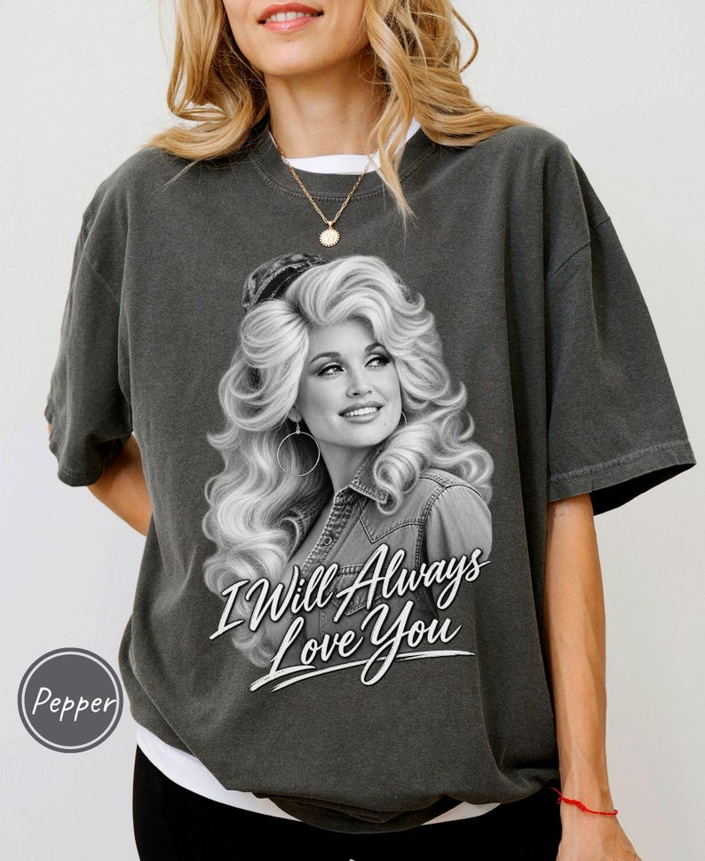 May include: A charcoal gray short-sleeve t-shirt featuring a black and white graphic of a woman with voluminous blonde hair wearing a denim shirt. The text "I Will Always Love You" is printed in white script below the portrait.
