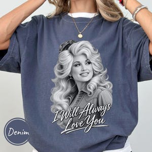 May include: A denim-colored crewneck t-shirt featuring a black and white graphic print of a woman with voluminous hair wearing a collared shirt. Below the portrait, the text reads I Will Always Love You in a white script font with a black outline.