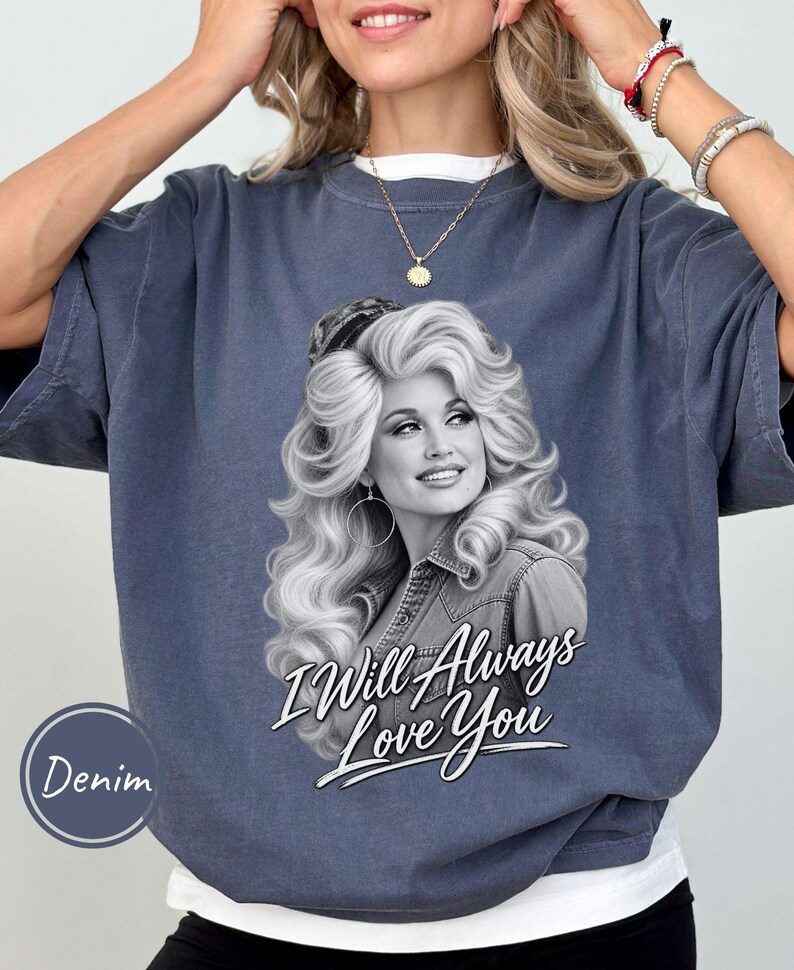 May include: A denim-colored crewneck t-shirt featuring a black and white graphic print of a woman with voluminous hair wearing a collared shirt. Below the portrait, the text reads I Will Always Love You in a white script font with a black outline.