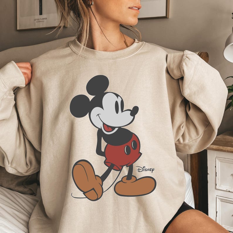 May include: Beige sweatshirt featuring a large graphic of Mickey Mouse in his classic pose. Mickey is depicted with black ears, a gray face, red shorts, and brown shoes. The Disney logo is printed on the lower right.