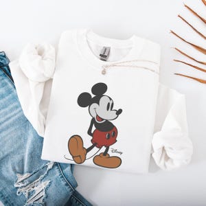 May include: White sweatshirt featuring a classic Mickey Mouse graphic. The cartoon character is depicted in his iconic red shorts, brown shoes, and black ears. The Disney logo is printed below the character.