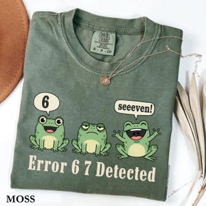 May include: A moss green t-shirt with a graphic of three cartoon frogs and the text "Error 6 7 Detected." The frogs have speech bubbles with the numbers "6" and the word "seeeven!" A silver necklace with a pendant is on the shirt.