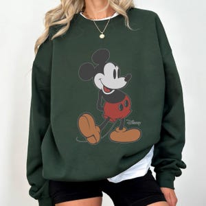 May include: Dark green sweatshirt featuring a classic Mickey Mouse graphic. The cartoon character is depicted in his iconic red shorts, brown shoes, and black ears. The Disney logo is visible on the bottom right.