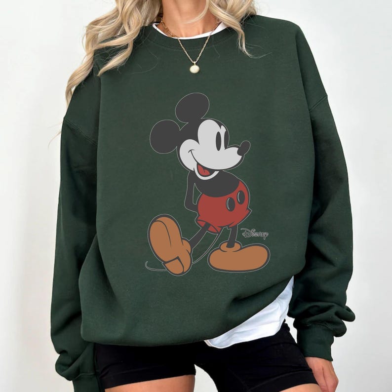 May include: Dark green sweatshirt featuring a classic Mickey Mouse graphic. The cartoon character is depicted in his iconic red shorts, brown shoes, and black ears. The Disney logo is visible on the bottom right.