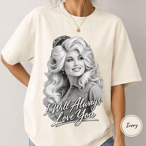 May include: An ivory short-sleeved t-shirt featuring a black and white graphic print of a woman with voluminous blonde hair wearing a denim shirt. The text "I Will Always Love You" is printed in a stylized black script font below the portrait.