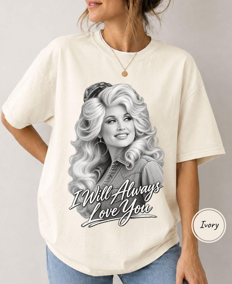 May include: An ivory short-sleeved t-shirt featuring a black and white graphic print of a woman with voluminous blonde hair wearing a denim shirt. The text "I Will Always Love You" is printed in a stylized black script font below the portrait.