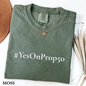May include: A moss green t-shirt with the text "#YesOnProp50" in white. The shirt has a crew neck and a small tag on the collar. A gold-colored necklace with a pendant is draped across the shirt. The word "MOSS" is at the bottom.
