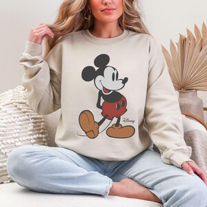 May include: Beige sweatshirt featuring a classic Mickey Mouse graphic. The cartoon character is depicted in black, red, and brown, with the Disney logo printed below. The sweatshirt has long sleeves and a crew neck.