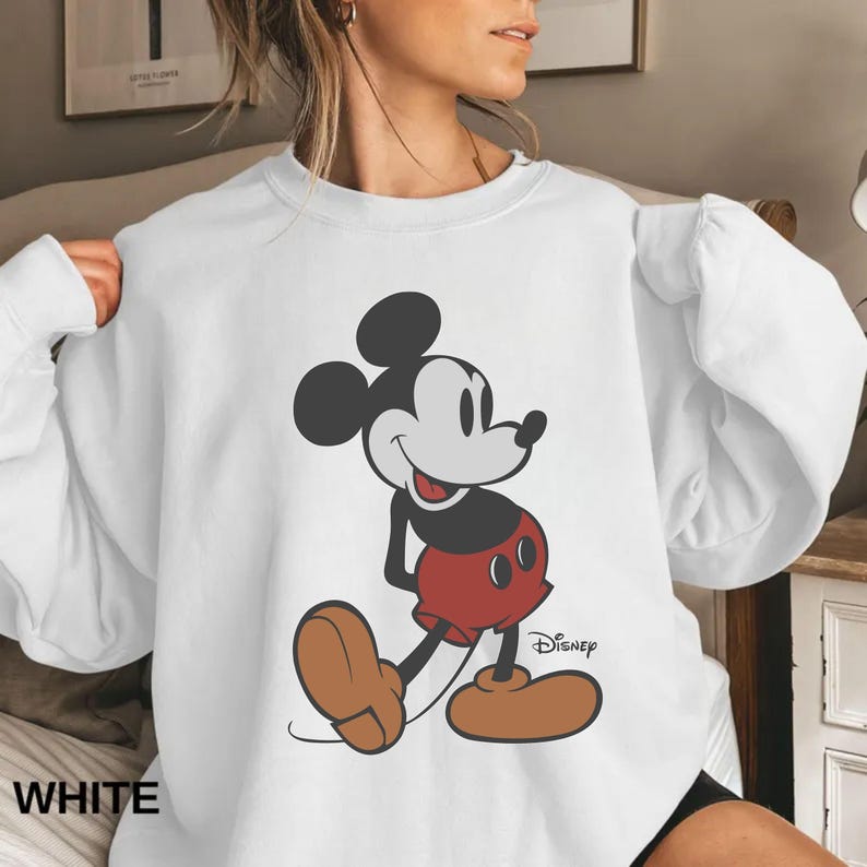 May include: White sweatshirt featuring a classic Mickey Mouse graphic. The cartoon character is depicted in black, red, and brown, with the Disney logo visible. The word "WHITE" is printed at the bottom left.