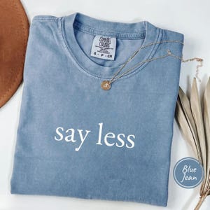 May include: A blue jean colored short-sleeve t-shirt with the text "say less" printed in white on the front. The shirt features a Comfort Colors brand label inside the collar and is paired with a thin gold-toned necklace with a round pendant.