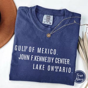 May include: A navy blue short-sleeve t-shirt with a crew neckline and a white graphic print. The text reads GULF OF MEXICO. JOHN F. KENNEDY CENTER. LAKE ONTARIO. The shirt features a Comfort Colors brand label at the inner neck and a size S tag.