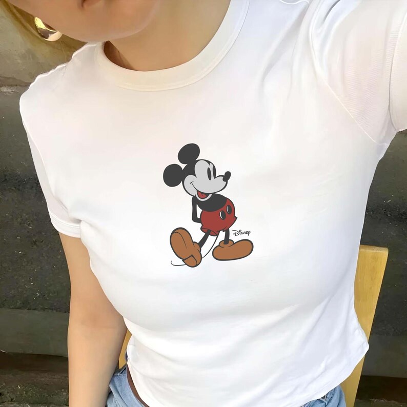 Puede incluir: Camiseta blanca con un gr&aacute;fico de Mickey Mouse. El personaje de dibujos animados se muestra con pantalones cortos rojos, zapatos marrones y orejas negras. El logotipo de Disney es visible en la parte inferior derecha.