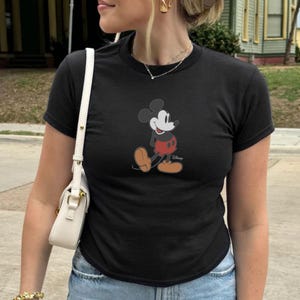 Puede incluir: Camiseta negra con un dibujo de Mickey Mouse. El personaje de dibujos animados se muestra en su pose cl&aacute;sica, con pantalones cortos rojos, zapatos amarillos y guantes blancos. La camiseta es de manga corta y tiene cuello redondo.