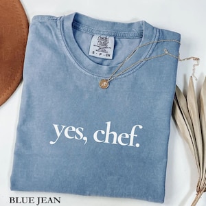 May include: A blue jean Comfort Colors t-shirt with the text "yes, chef." A gold necklace with a pendant is draped over the shirt. The shirt is folded and has a tag that reads "Comfort Colors."