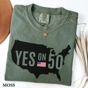 May include: A moss green t-shirt with a black outline of the United States and the text "YES ON 50" in gray. The American flag is featured below the text. A gold necklace with a pendant is draped on the shirt.