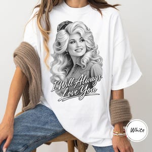 May include: A white t-shirt featuring a black and white graphic print of a woman with voluminous blonde hair, wearing hoop earrings and a denim shirt. Below the portrait, the text reads "I Will Always Love You" in a stylized script font.
