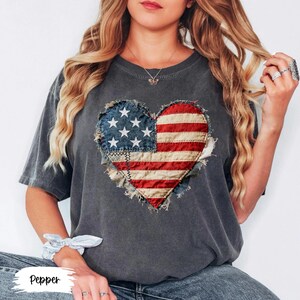 May include: A dark gray t-shirt featuring a heart-shaped design of the American flag. The flag design is made of patches of blue, red, and white fabric. The word "Pepper" is printed at the bottom of the image.