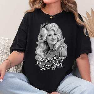 May include: A black short-sleeved t-shirt featuring a grayscale graphic print of a woman with voluminous blonde hair wearing a denim shirt. Below the portrait, the white cursive text reads I Will Always Love You, centered on the chest.
