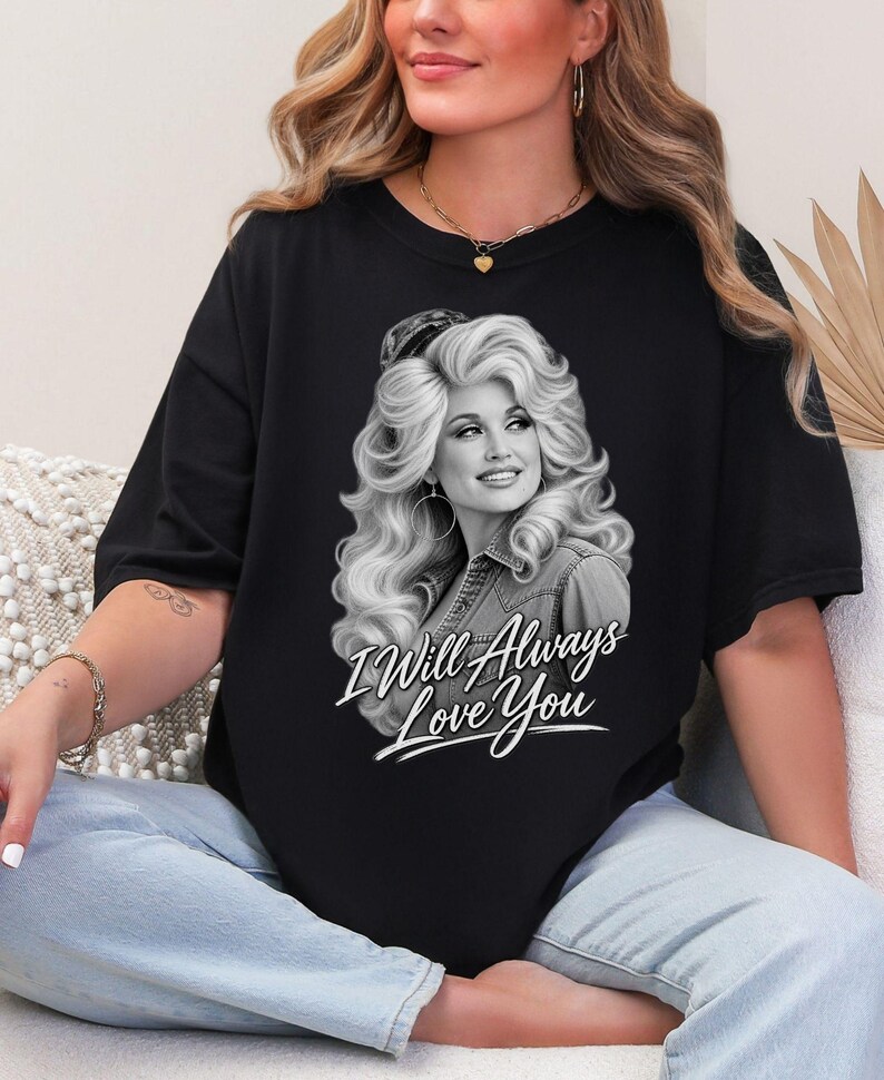 May include: A black short-sleeved t-shirt featuring a grayscale graphic print of a woman with voluminous blonde hair wearing a denim shirt. Below the portrait, the white cursive text reads I Will Always Love You, centered on the chest.
