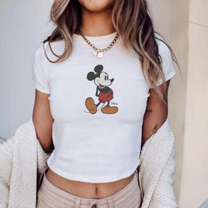 Puede incluir: Top corto blanco con un gr&aacute;fico de Mickey Mouse. El personaje de dibujos animados se muestra con pantalones cortos rojos, zapatos marrones y orejas negras. Una prenda casual para el d&iacute;a a d&iacute;a. El logotipo de Disney es visible.