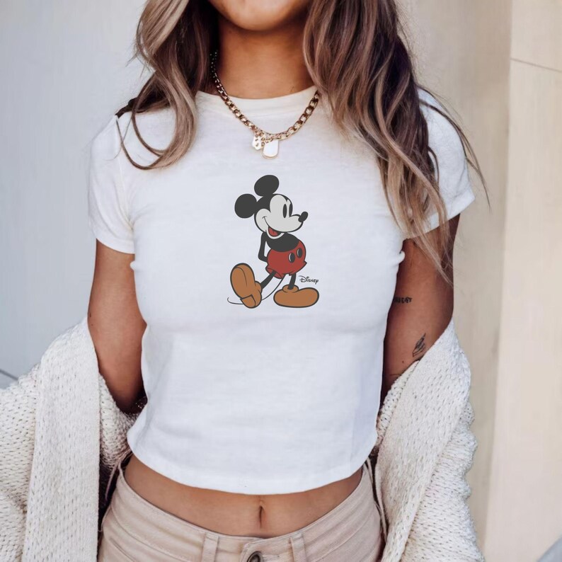 Puede incluir: Top corto blanco con un gr&aacute;fico de Mickey Mouse. El personaje de dibujos animados se muestra con pantalones cortos rojos, zapatos marrones y orejas negras. Una prenda casual para el d&iacute;a a d&iacute;a. El logotipo de Disney es visible.