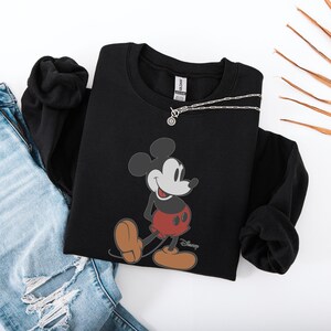 May include: Black crewneck sweatshirt featuring a smiling Mickey Mouse graphic in red, black, and tan. A silver necklace with a circular pendant rests on the sweatshirt. The sweatshirt is paired with blue jeans.