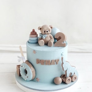 Pu&ograve; includere: Una torta di compleanno azzurra decorata con un orsacchiotto, il numero 2 e il nome "DEHAT". La torta presenta anche un elefante giocattolo su ruote, una ciambella e una pila di anelli. La torta &egrave; su un supporto bianco.