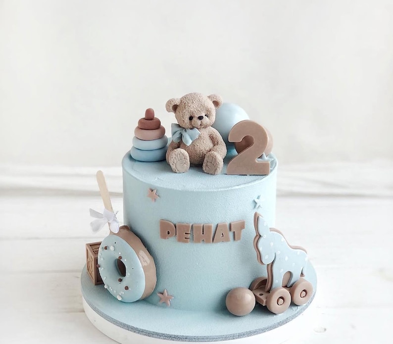 Pu&ograve; includere: Una torta di compleanno azzurra decorata con un orsacchiotto, il numero 2 e il nome "DEHAT". La torta presenta anche un elefante giocattolo su ruote, una ciambella e una pila di anelli. La torta &egrave; su un supporto bianco.