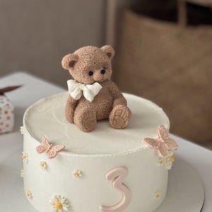 Pu&ograve; includere: Una torta bianca glassata decorata con un orsacchiotto marrone con un papillon bianco. La torta &egrave; adornata con farfalle rosa, piccoli fiori bianchi e il numero 3 in rosa. La torta &egrave; su un vassoio bianco.
