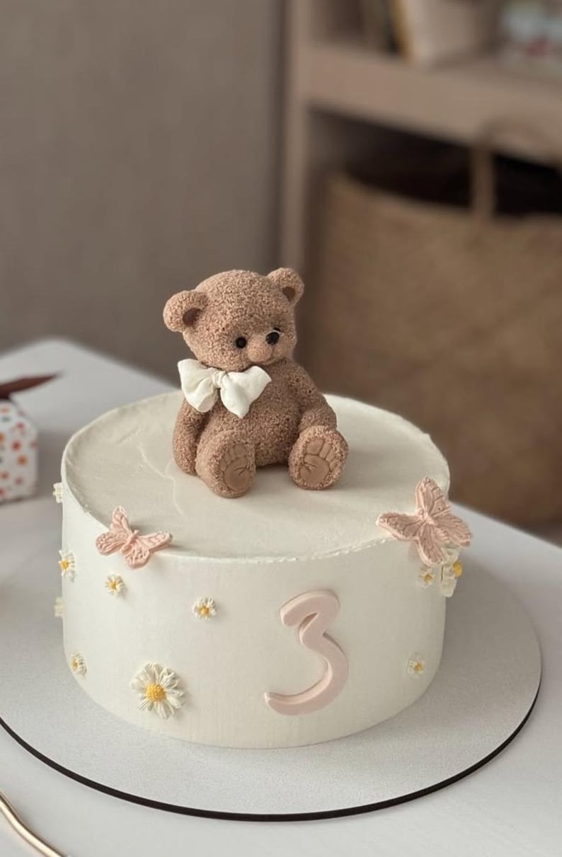 Pu&ograve; includere: Una torta bianca glassata decorata con un orsacchiotto marrone con un papillon bianco. La torta &egrave; adornata con farfalle rosa, piccoli fiori bianchi e il numero 3 in rosa. La torta &egrave; su un vassoio bianco.
