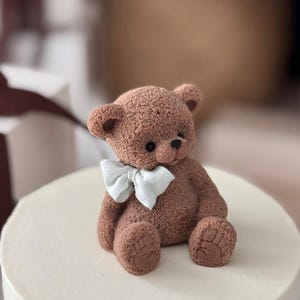 Pu&ograve; includere: Un orsetto di peluche marrone con un papillon bianco &egrave; posto sopra una torta bianca glassata. L'orso ha zampe e orecchie dettagliate. La torta &egrave; rotonda e sembra un dessert.