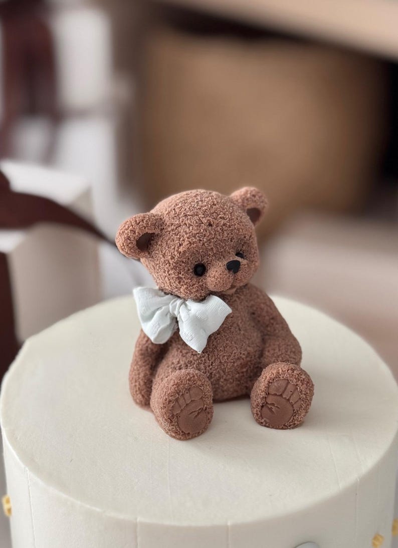 Pu&ograve; includere: Un orsetto di peluche marrone con un papillon bianco &egrave; posto sopra una torta bianca glassata. L'orso ha zampe e orecchie dettagliate. La torta &egrave; rotonda e sembra un dessert.
