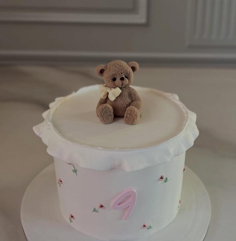 Pu&ograve; includere: Una torta bianca con un orsetto in cima e il numero 9 rosa. La torta ha un bordo a balze ed &egrave; decorata con piccoli fiori rosa e foglie verdi. L'orsetto &egrave; marrone con un fiocco color crema.