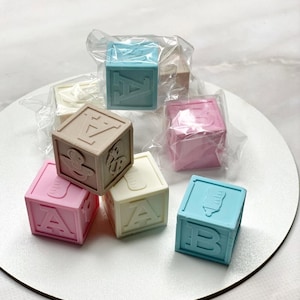 Puede incluir: Un conjunto de bloques de bebé coloridos en tonos rosa, azul, beige y blanco. Cada bloque presenta una letra o un icono relacionado con bebés. Los bloques están envueltos individualmente en plástico transparente.