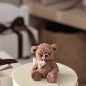 Pu&ograve; includere: Un orsacchiotto marrone con un fiocco rosa &egrave; seduto su una torta bianca. L'orso ha dettagli e &egrave; circondato da scatole regalo bianche con nastri marroni. La torta ha un bordo rosa chiaro.