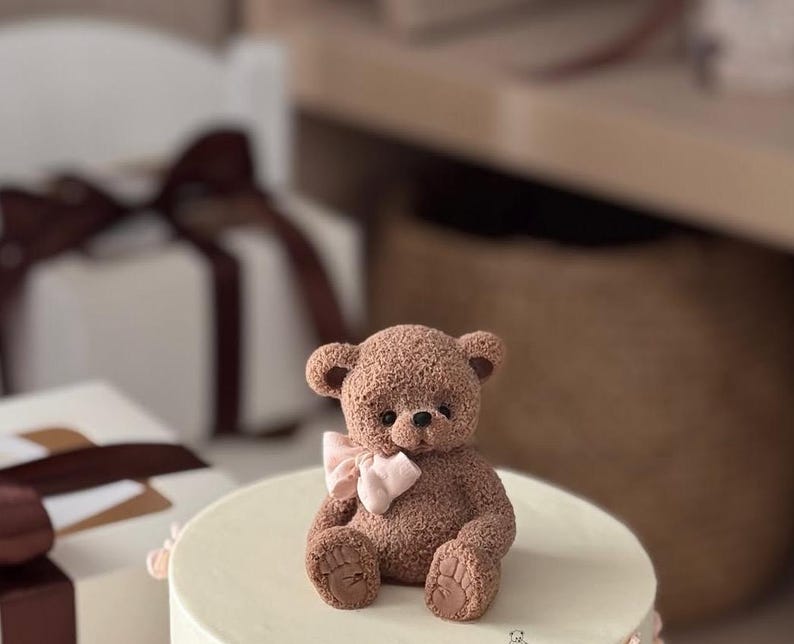 Pu&ograve; includere: Un orsacchiotto marrone con un fiocco rosa &egrave; seduto su una torta bianca. L'orso ha dettagli e &egrave; circondato da scatole regalo bianche con nastri marroni. La torta ha un bordo rosa chiaro.