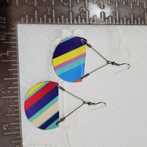 May include: A pair of colorful striped earrings with silver hooks. The earrings are made of metal and have a semi-circular shape. The stripes are red, yellow, blue, green, and purple.