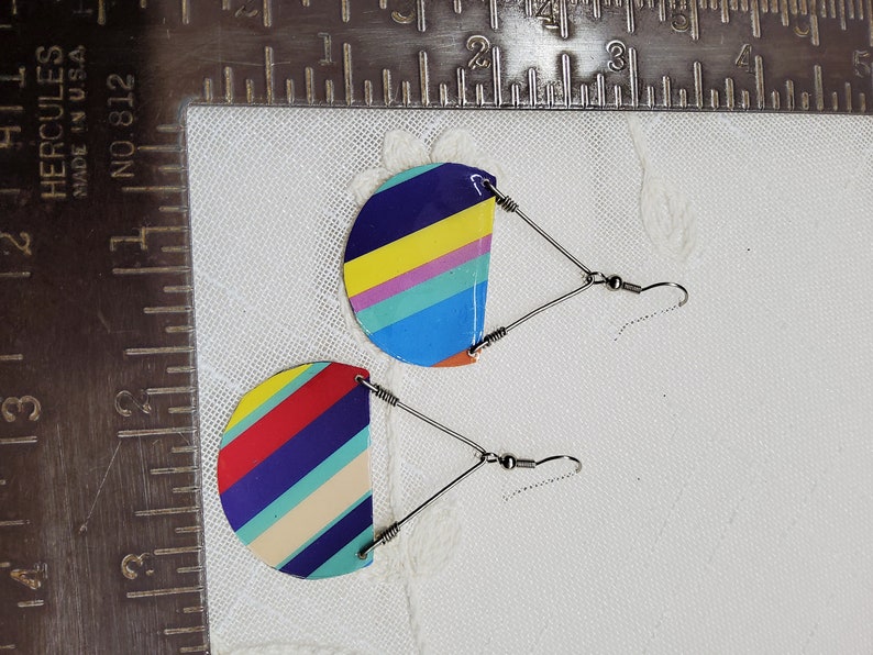 May include: A pair of colorful striped earrings with silver hooks. The earrings are made of metal and have a semi-circular shape. The stripes are red, yellow, blue, green, and purple.