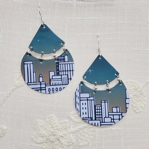 May include: A pair of dangle earrings featuring a cityscape design. The earrings are made of metal and have a teal and white color scheme.