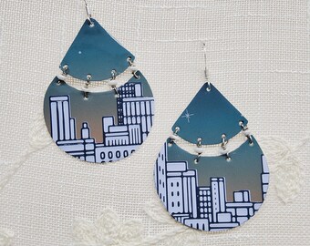 May include: A pair of dangle earrings featuring a cityscape design. The earrings are made of metal and have a teal and white color scheme.