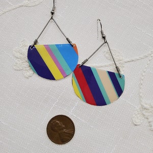 May include: A pair of colorful striped earrings with a half-circle shape. The earrings are made of metal and have a silver finish. The stripes are in shades of blue, yellow, red, pink, and white.
