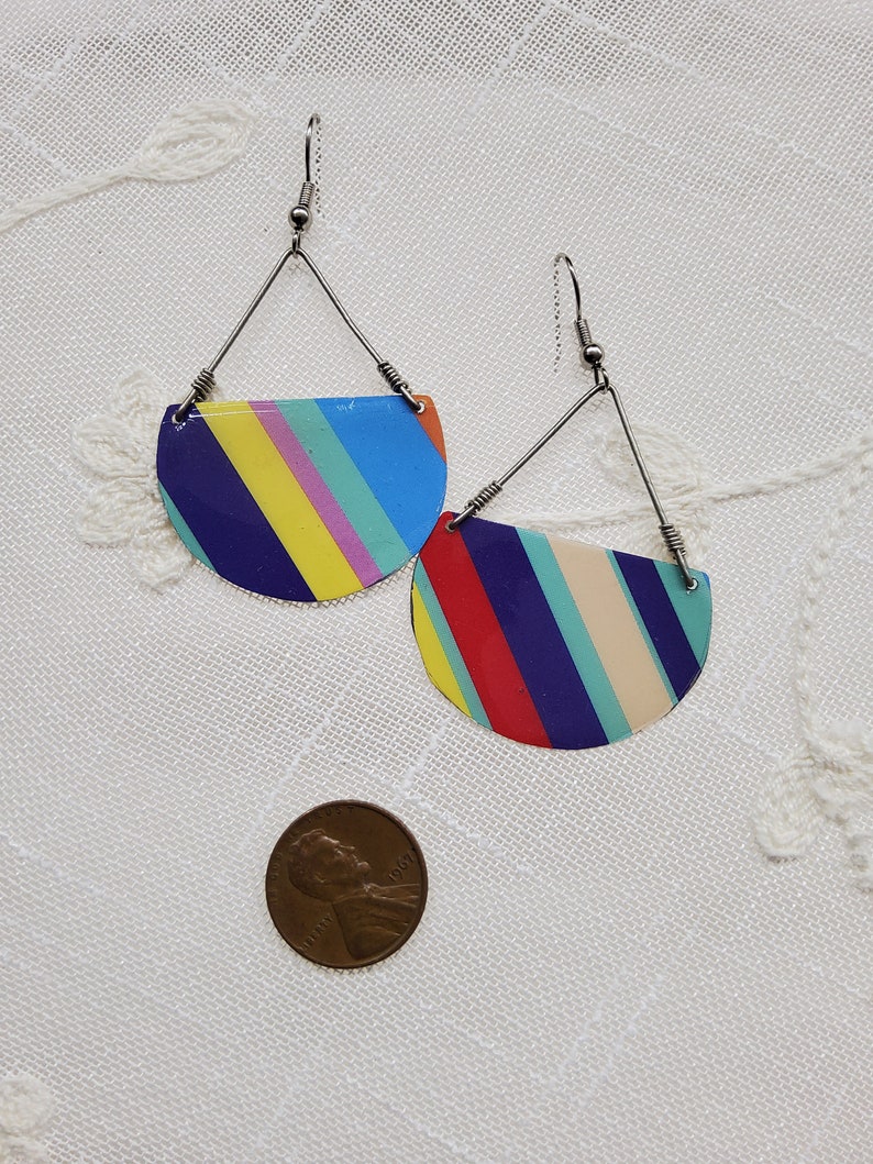 May include: A pair of colorful striped earrings with a half-circle shape. The earrings are made of metal and have a silver finish. The stripes are in shades of blue, yellow, red, pink, and white.