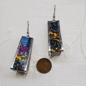 May include: A pair of silver earrings with a rectangular shape. Each earring features a colorful abstract painting inside. The earrings are about the size of a US penny.