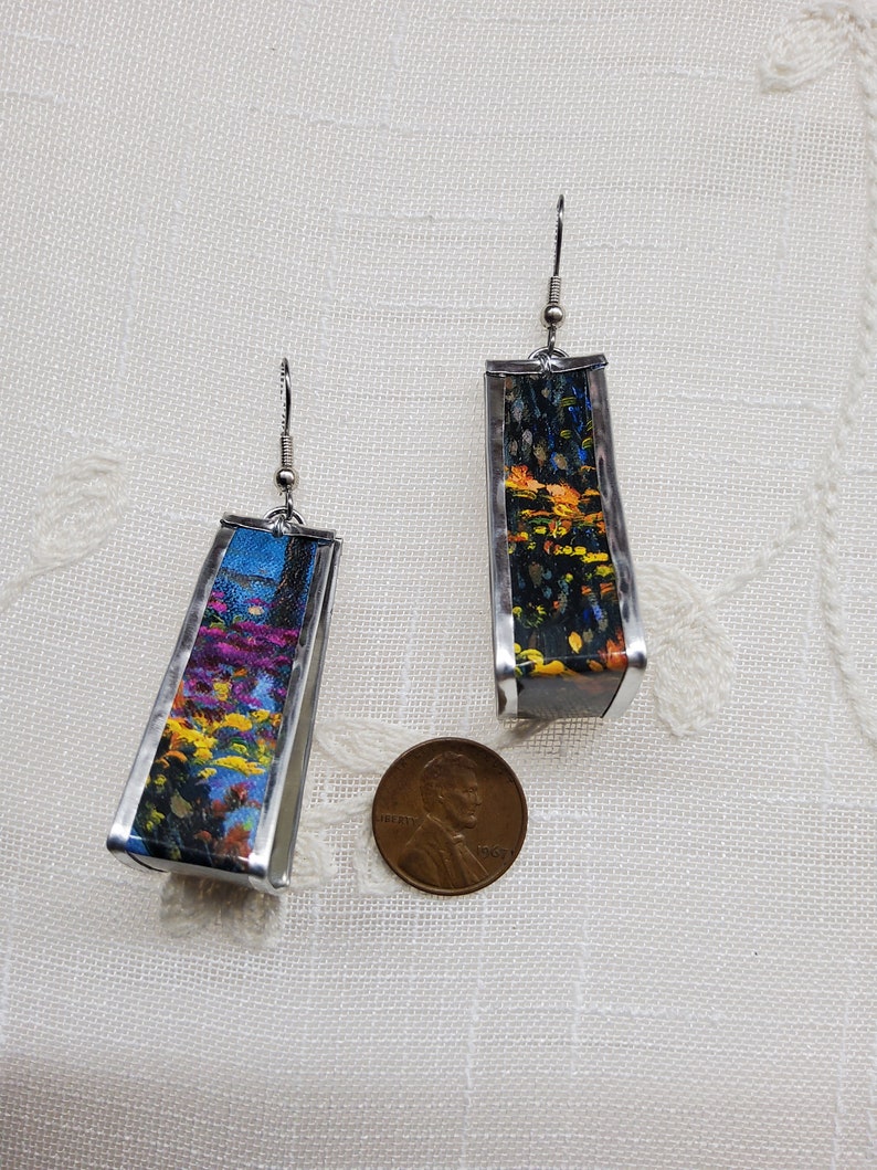 May include: A pair of silver earrings with a rectangular shape. Each earring features a colorful abstract painting inside. The earrings are about the size of a US penny.