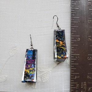 May include: A pair of silver earrings with a rectangular design. Each earring features a colorful abstract image with shades of blue, yellow, orange, and black.
