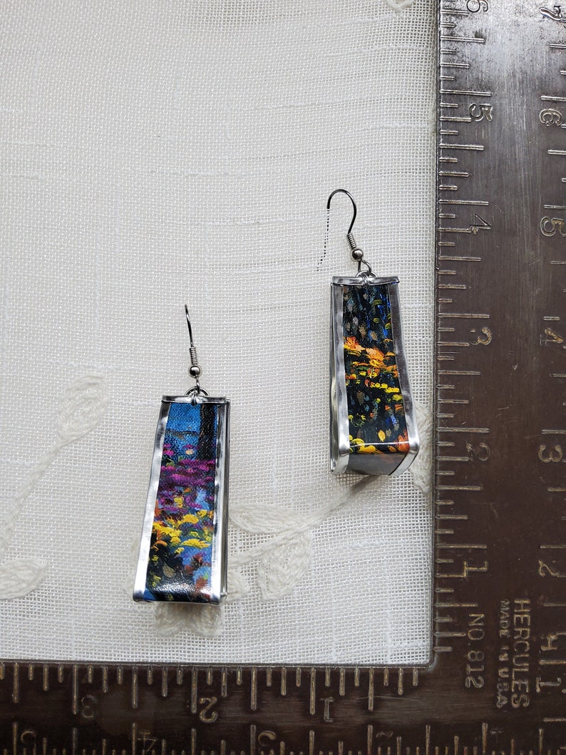 May include: A pair of silver earrings with a rectangular design. Each earring features a colorful abstract image with shades of blue, yellow, orange, and black.